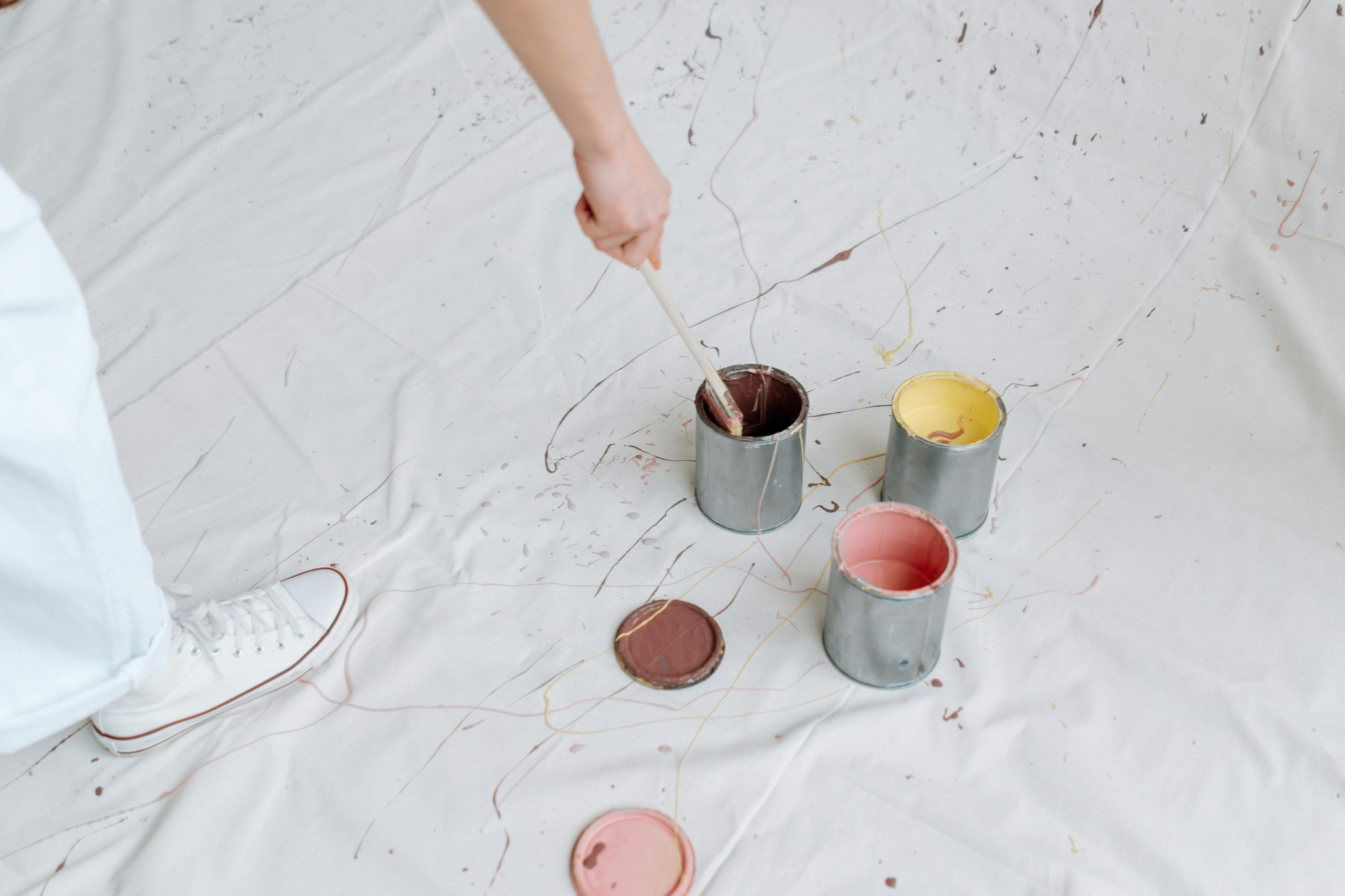 Close-up of paint cans and brushes on a white fabric backdrop, creating a messy artistic scene. Gros plan de pots de peinture et de pinceaux sur un fond de tissu blanc, créant une scène artistique désordonnée.