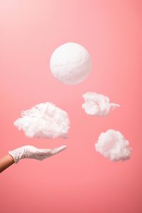 An artistic composition of floating clouds and hand with medical glove against a pink backdrop. Une composition artistique de nuages flottants et d'une main avec un gant médical sur un fond rose.