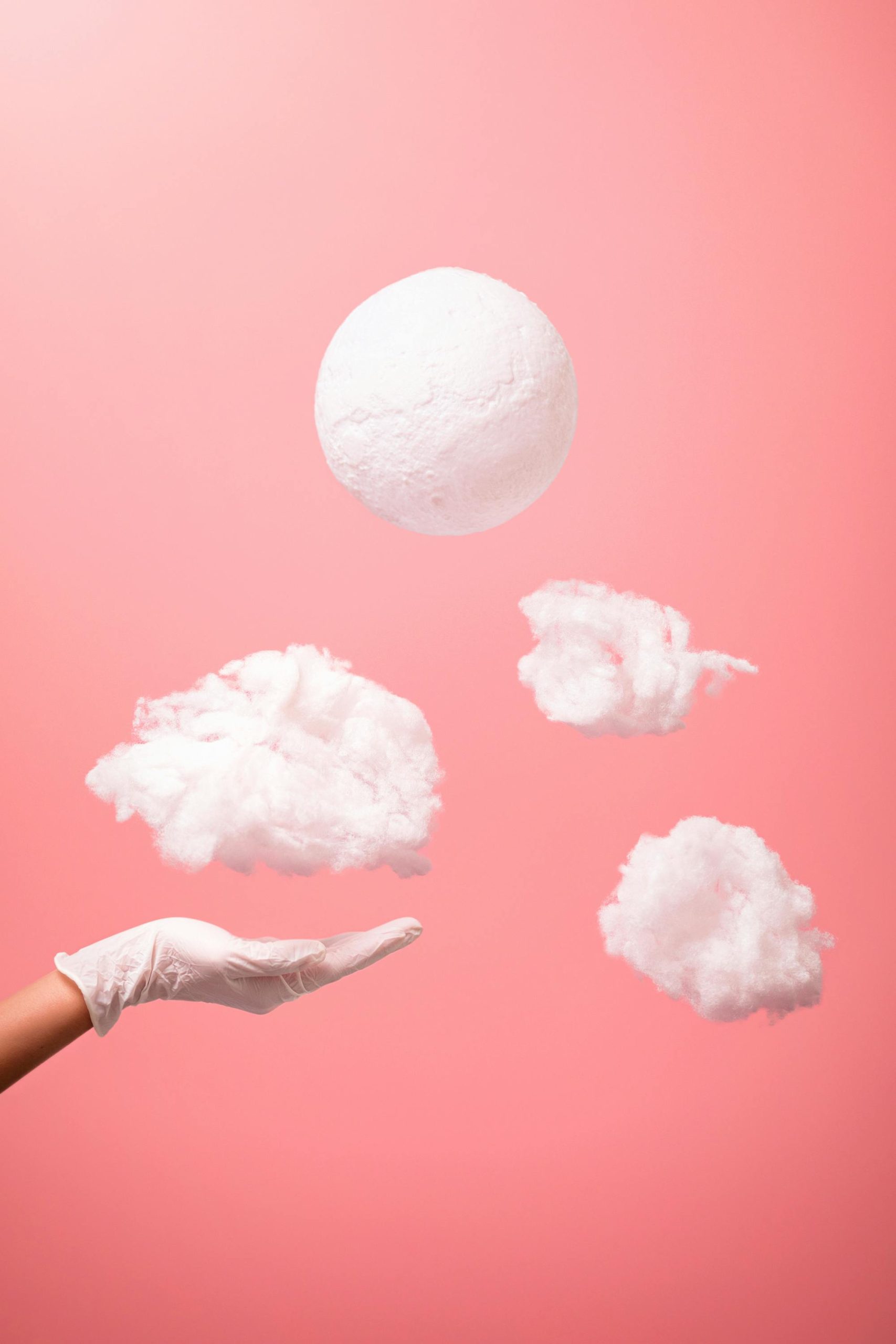 An artistic composition of floating clouds and hand with medical glove against a pink backdrop. Une composition artistique de nuages flottants et d'une main avec un gant médical sur un fond rose.