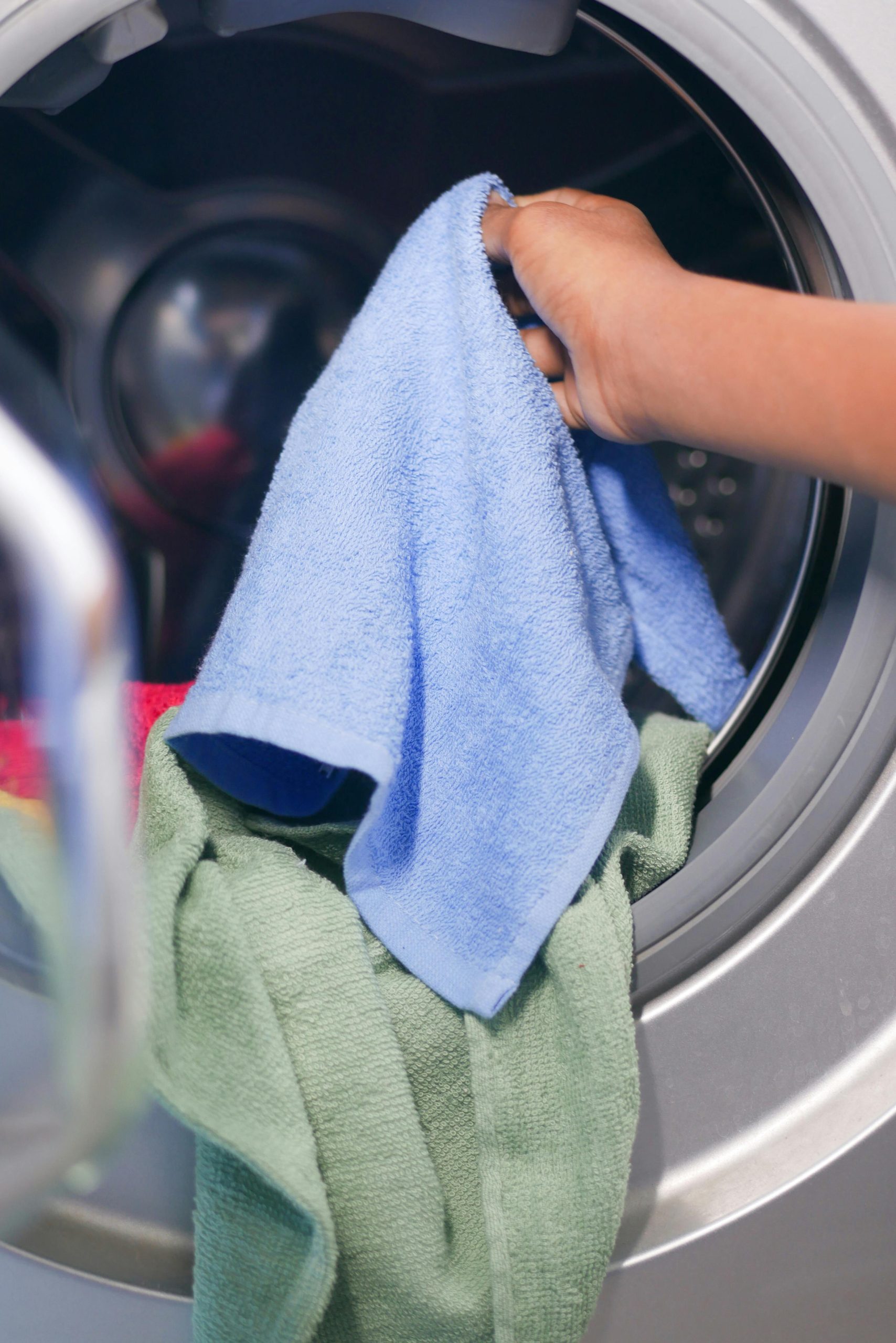 Close-up of a hand placing colorful towels into a front-loading washing machine. Gros plan d'une main plaçant des serviettes colorées dans une machine à laver à chargement frontal.