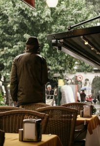 A man stands in a rainy outdoor café with empty wicker chairs and tables. Un homme se tient dans un café extérieur pluvieux avec des chaises et des tables en osier vides.