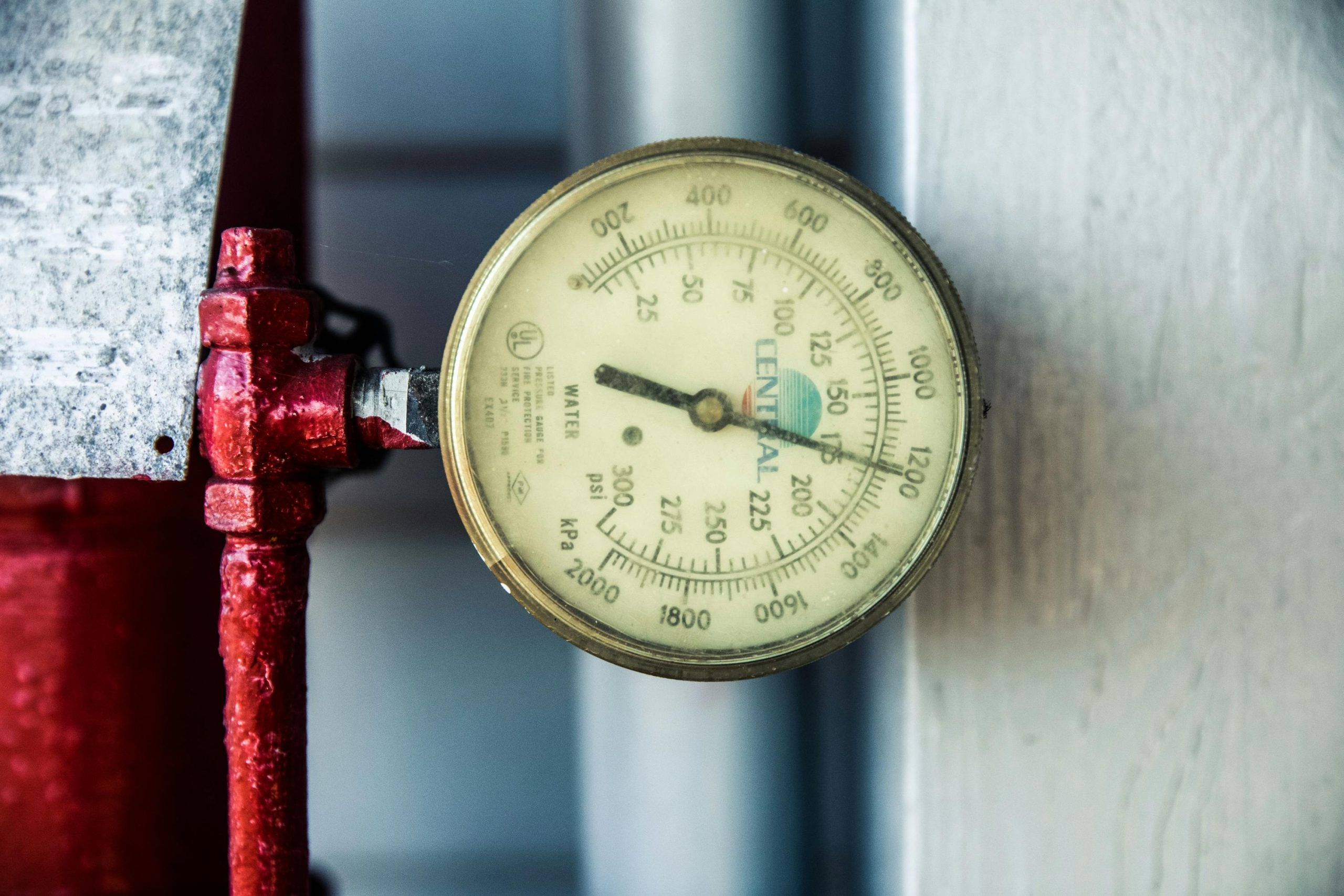 Detailed view of a pressure gauge attached to a red industrial pipe, measuring PSI and kPa. Vue détaillée d'un manomètre attaché à un tuyau industriel rouge, mesurant PSI et kPa.
