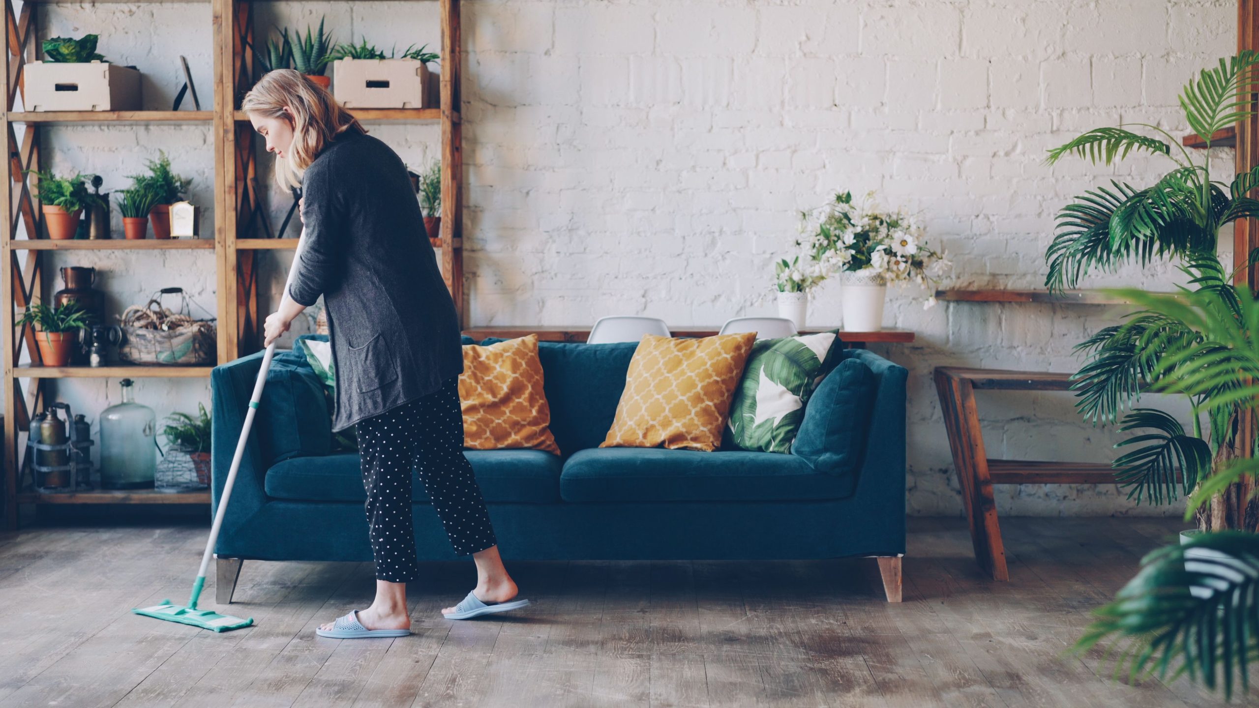 Woman with mop cleaning cozy living room with blue sofa and plants. Femme avec un balai-serpillière nettoyant un salon confortable avec canapé bleu et plantes.