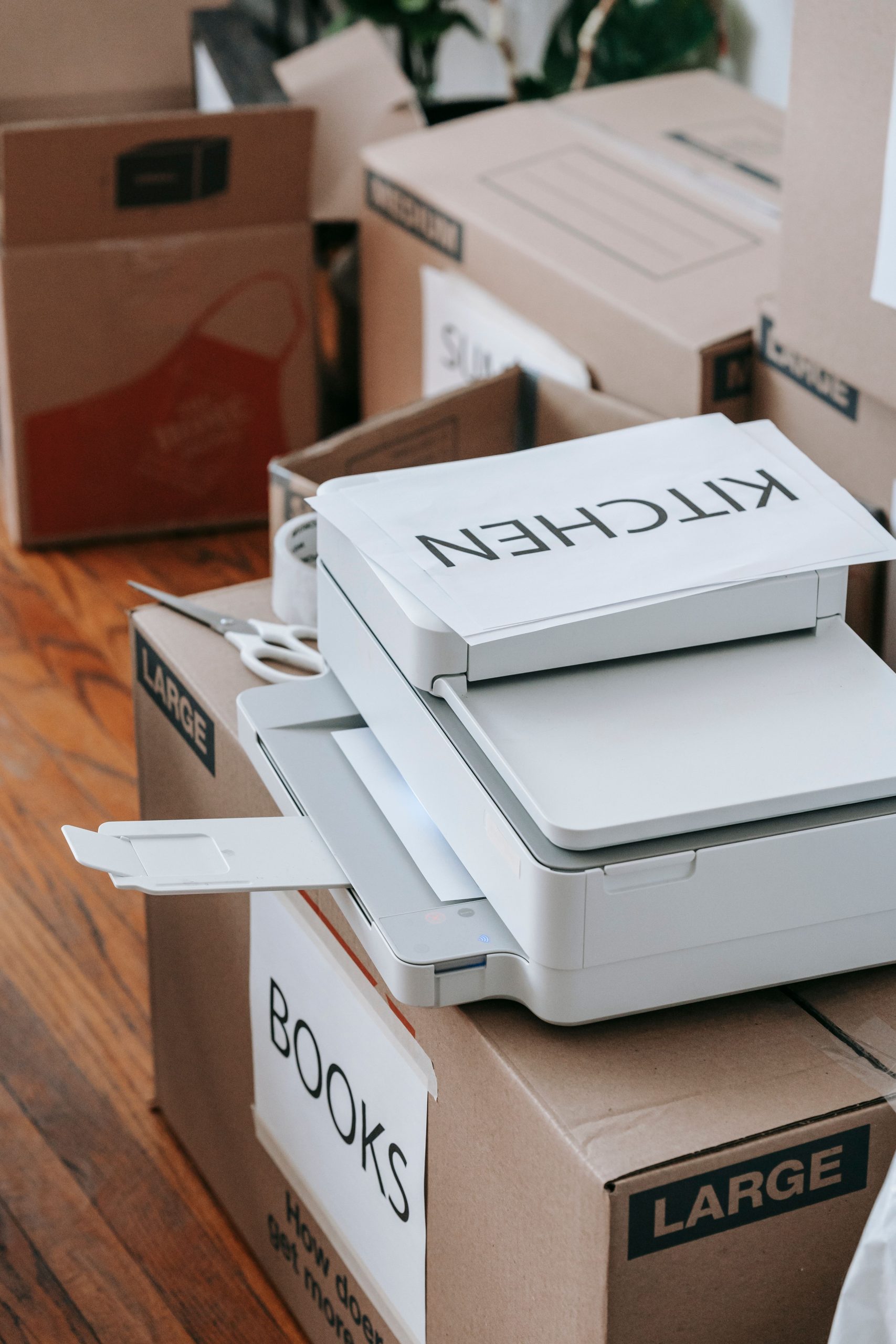 A collection of large cardboard boxes labeled for moving, alongside a printer, representing organization and preparation indoors. Une collection de grandes boîtes en carton étiquetées pour le déménagement, aux côtés d'une imprimante, représentant l'organisation et la préparation en intérieur.